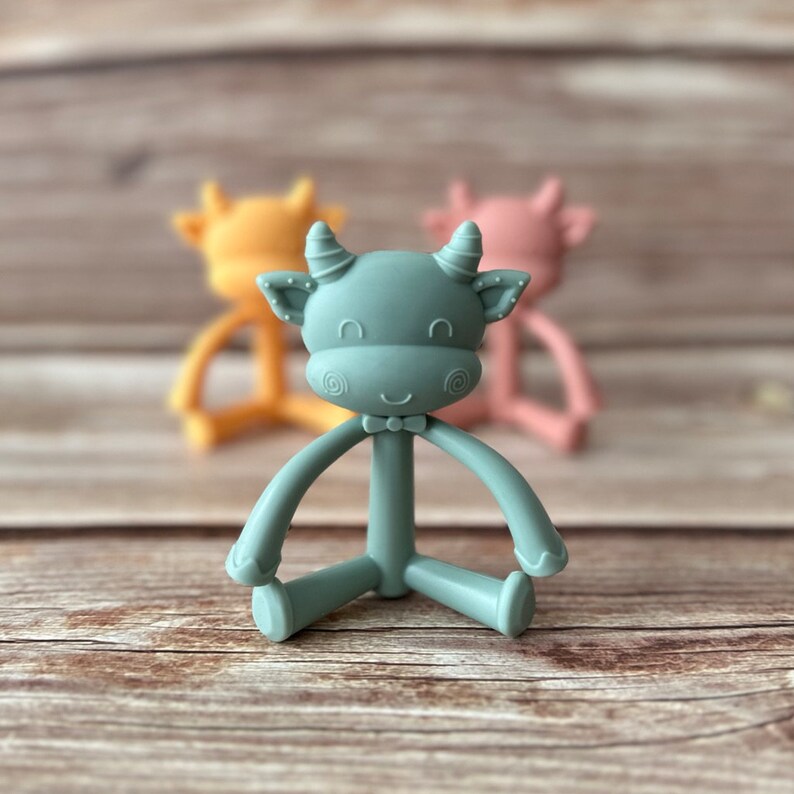 May include: A teal blue silicone baby teether shaped like a cow with a bow tie. The teether has a textured surface and is designed for babies to chew on.