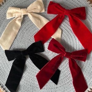 May include: Four velvet bows in cream, red, black, and burgundy are displayed on a grey, textured surface. Each bow features long, flowing tails. These bows are likely hair accessories or decorative items.