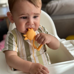 May include: A baby in a striped shirt is chewing on a yellow silicone teether shaped like a rhinoceros.
