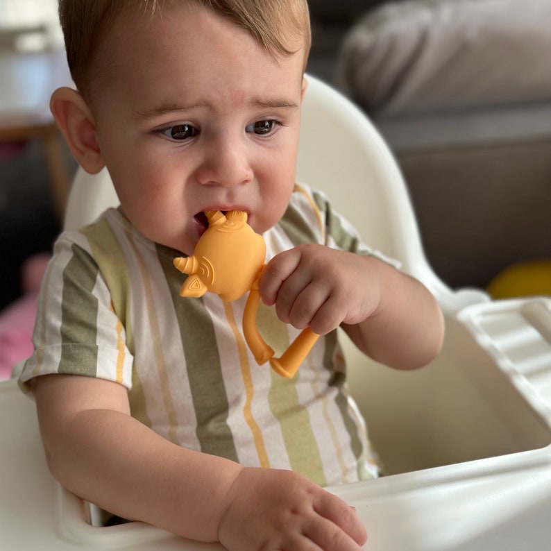 May include: A baby in a striped shirt is chewing on a yellow silicone teether shaped like a rhinoceros.