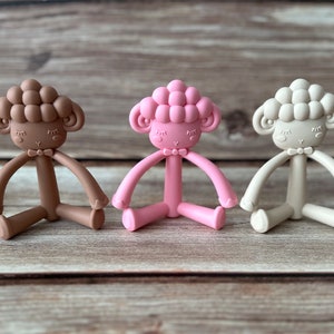 May include: Three silicone baby teethers shaped like sheep in brown, pink, and beige colors. Each teether has a bow tie and a cute face.