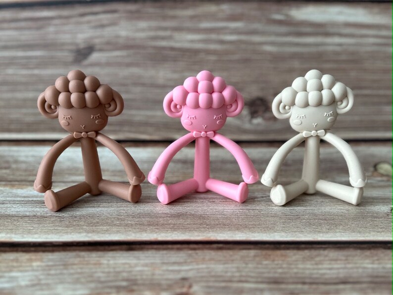 May include: Three silicone baby teethers shaped like sheep in brown, pink, and beige colors. Each teether has a bow tie and a cute face.