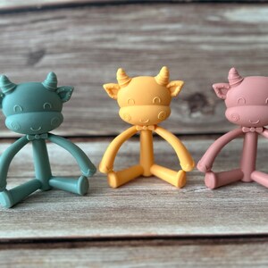 May include: Three silicone baby teethers shaped like cows in green, yellow, and pink. Each teether has a bow tie and a smiling face.