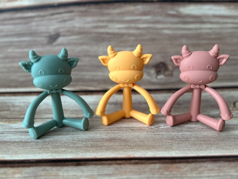 May include: Three silicone baby teethers shaped like cows in green, yellow, and pink. Each teether has a bow tie and a smiling face.