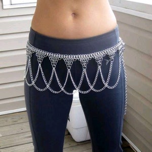 May include: A silver chainmail belt with a dangling chain fringe. The belt is worn over black pants.