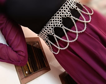 May include: A silver chainmail belt with a decorative design. The belt is worn over a black top and a burgundy skirt.
