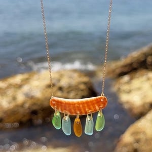 Peut inclure: Un collier à chaîne dorée fine avec un pendentif en coquillage incurvé rayé d'orange. Cinq petits morceaux de verre de mer lisses et translucides, dans des tons vert clair, bleu et ambre, sont suspendus au coquillage par des anneaux dorés.