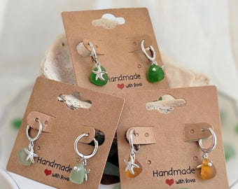 Puede incluir: Se muestran tres pares de pendientes hechos a mano. Cada par tiene un aro o gancho plateado con un dije. Los dijes incluyen una estrella de mar y vidrio de mar en tonos verdes y naranjas. Los pendientes se exhiben en etiquetas de cartón marrón.