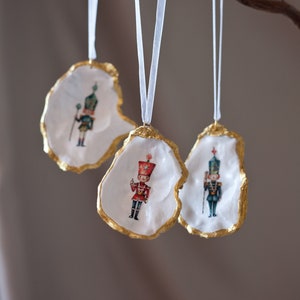 Puede incluir: Tres adornos de concha de ostra blanca con ribete dorado, cada uno con una ilustración vintage diferente de un soldado de juguete. Los adornos cuelgan de una cinta blanca en una rama.