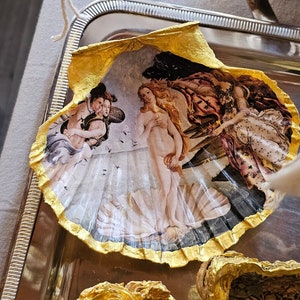 Può includere: Una conchiglia con un bordo dorato, che presenta un dipinto della Nascita di Venere, una famosa opera d'arte rinascimentale. Il dipinto raffigura una donna nuda in piedi su una conchiglia, con due figure che la fiancheggiano.