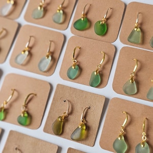 Puede incluir: Una colección de pendientes de vidrio de mar expuestos en tarjetas. Los pendientes están hechos con ganchos de color dorado y presentan varios tonos de vidrio de mar verde.