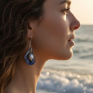 Pu&ograve; includere: Una donna indossa orecchini pendenti. Ogni orecchino presenta un elemento a forma di conchiglia blu e bianca con una perla pendente. Lo sfondo mostra l'oceano e un tramonto.