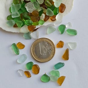 May include: A collection of small, colourful sea glass pieces in shades of green, brown, and white. The sea glass is arranged around a Euro coin for scale.