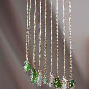 K&ouml;nnte beinhalten: Ein Set aus neun goldenen Kettenhalsketten mit verschiedenen farbigen Glasanh&auml;ngern. Die Anh&auml;nger sind in einem goldenen Drahtk&auml;figdesign eingefasst. Die Farben des Glases sind gr&uuml;n und blau.