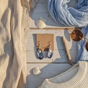 Pu&ograve; includere: Una composizione a tema spiaggia su una superficie di legno bianco. Include una camicia beige, una sciarpa azzurra, un maglione bianco, occhiali da sole e orecchini con dettagli blu. Sono presenti anche conchiglie.