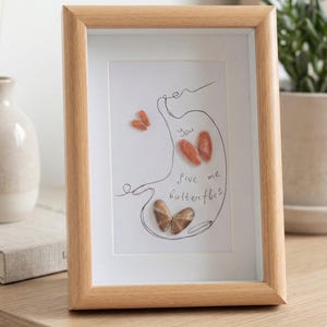 Può includere: Un'opera d'arte incorniciata con una cornice in legno chiaro. L'opera presenta un disegno a tratto di una forma di stomaco con le parole "You give me butterflies" e diverse forme di farfalle a forma di conchiglia.