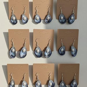 Pu&ograve; includere: Nove paia di orecchini pendenti esposti su cartoncini marroni. Ogni orecchino presenta un design a conchiglia blu e argento con una perla bianca. Gli orecchini hanno ganci argentati.