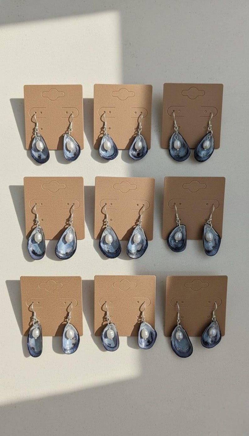 Pu&ograve; includere: Nove paia di orecchini pendenti esposti su cartoncini marroni. Ogni orecchino presenta un design a conchiglia blu e argento con una perla bianca. Gli orecchini hanno ganci argentati.
