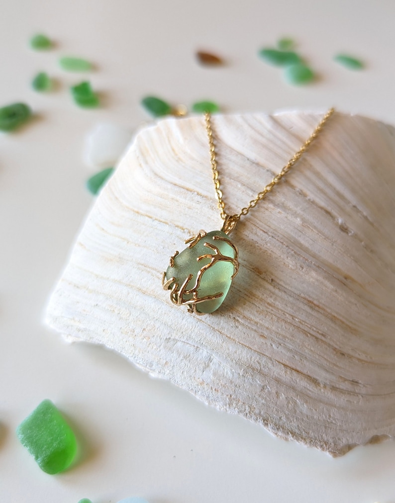 K&ouml;nnte beinhalten: Eine goldene Kettenkette mit einem gr&uuml;nen Meerglass Anh&auml;nger, der in einem filigranen Golddrahtdesign eingefasst ist. Die Kette wird auf einer wei&szlig;en Muschel pr&auml;sentiert.