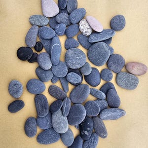 Può includere: Una collezione di pietre lisce, grigie e nere di varie dimensioni. Le pietre sono disposte in uno schema casuale su una superficie marrone chiaro.