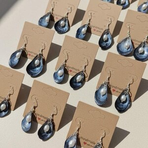 Pu&ograve; includere: Orecchini fatti a mano con un design a conchiglia blu e bianca e una perla pendente. Gli orecchini sono esposti su piccoli cartellini di cartone marrone. Gli orecchini hanno ganci color argento.