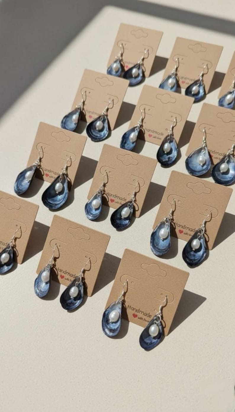 Pu&ograve; includere: Orecchini fatti a mano con un design a conchiglia blu e bianca e una perla pendente. Gli orecchini sono esposti su piccoli cartellini di cartone marrone. Gli orecchini hanno ganci color argento.