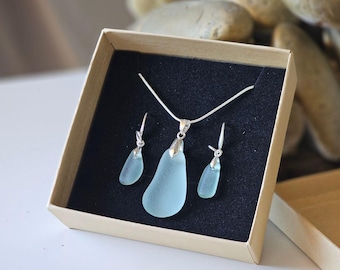 May include: A set of sea glass jewelry in a box. The set includes a necklace and earrings. The sea glass is a light blue color and the jewelry is made with silver.