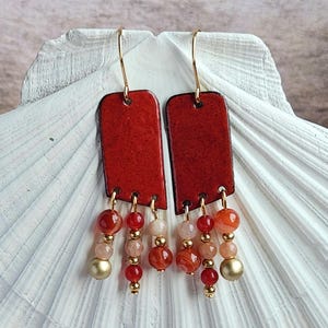 May include: Handmade earrings with red enamel rectangular pendants and gold-coloured hooks. The earrings have dangling strands of red, orange, and pink beads, with gold-coloured accents and a gold-coloured bead at the bottom.