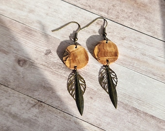 May include: A pair of dangle earrings featuring round, light brown wooden discs with a natural grain pattern. Below each disc hangs a dark bronze-colored feather-shaped charm. The earrings have bronze-colored ear wires.