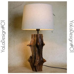 Puede incluir: Una lámpara de mesa con una pantalla de tela blanca y una base de madera que se asemeja a un tronco de árbol. La lámpara está encendida y proyecta un brillo cálido.