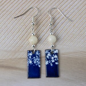 Puede incluir: Un par de pendientes colgantes. Cada pendiente presenta un colgante rectangular de esmalte azul oscuro con motas blancas. Encima del colgante hay una cuenta de color crema y detalles metálicos plateados. Los pendientes tienen ganchos plateados.
