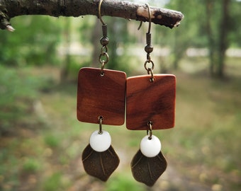 May include: A pair of wooden earrings with a brown square shape and a white ceramic leaf charm. The earrings are hanging from a branch.