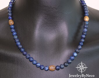 May include: A beaded necklace featuring small, round, deep blue beads. The necklace has three larger, decorative wooden beads with an engraved design. The necklace is displayed on a black mannequin.