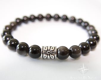 May include: A black beaded bracelet with a silver-colored cylindrical accent bead. The bracelet features polished, round black beads strung together. The accent bead has a floral design. The bracelet is a piece of jewelry.