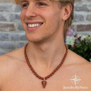 May include: A necklace featuring round, reddish-brown stone beads interspersed with small, flat, metallic gold-toned spacers. The piece includes a central, downward-pointing, triangular-shaped stone pendant in a matching reddish-brown hue.