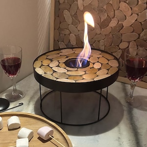 May include: A circular tabletop fire pit featuring a black metal frame and a top surface inlaid with smooth, tan-coloured river stones. A single flame rises from the central burner, flanked by two stemmed glasses filled with red wine.