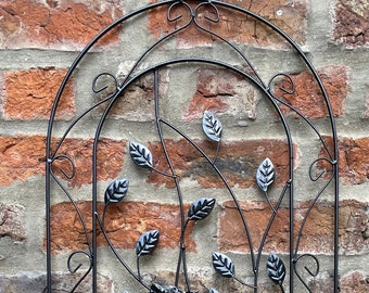May include: Black metal garden trellis featuring a vase and leaves design. The trellis is arched at the top and has a decorative scrollwork pattern.