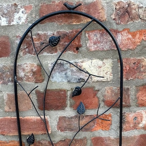 May include: Black metal garden trellis with a decorative design of leaves and a bird. The trellis is against a brick wall.