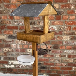 May include: A wooden bird feeder with a slate roof and a white ceramic bowl attached to a wooden pole. The feeder has a metal hook for hanging a suet feeder.