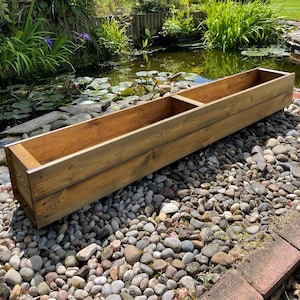 May include: A long, wooden planter box with two compartments. The planter is made of reclaimed wood and has a natural, rustic finish. It is sitting on a bed of small, grey stones.