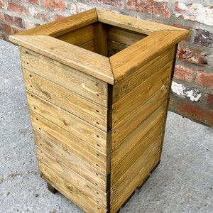 May include: A square wooden planter box with a natural wood finish. The planter has a wide opening and is made of reclaimed wood.