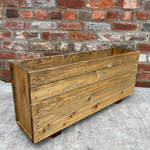 May include: A rectangular wooden planter box with a natural finish. The box is made from horizontal wooden planks and rests on small, dark brown feet. The background is a weathered brick wall.