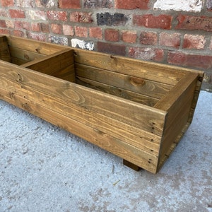 May include: A long, rectangular wooden planter box with two compartments. The planter is made of natural wood with a brown stain and has a slightly rounded edge. It sits on four small wooden legs.