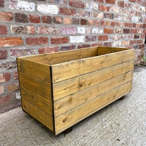 May include: A rectangular wooden planter box with a natural finish. The box is constructed from horizontal wooden planks and sits on small wooden feet. The background features a brick wall.