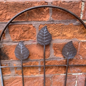 May include: A metal garden trellis with three leaf-shaped cut-outs. The trellis is brown and has a curved top. It is mounted on a brick wall.