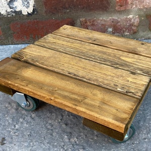 May include: A square wooden platform with four wheels, made from reclaimed wood with a dark brown stain. The platform is ideal for moving plants or other heavy objects.