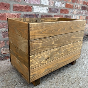 May include: A rectangular wooden planter box with a weathered, brown finish. The box is constructed from horizontal planks and sits on four small feet. The background features a brick wall.