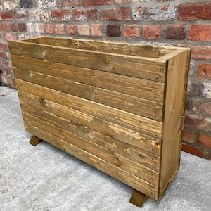 May include: A rustic wooden planter box with a natural brown finish. The planter has a rectangular shape and is made of reclaimed wood planks. It has a simple design with a slightly raised lip around the top and sits on four small legs.