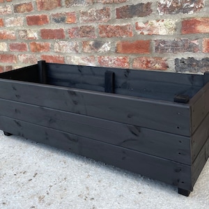 May include: A black wooden planter box with a rectangular shape and a simple design. The planter is made of dark wood planks and has a smooth finish.