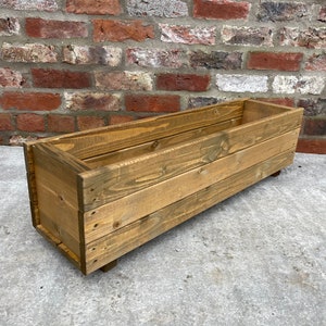 May include: A long, rectangular wooden planter box with a dark brown stain. The planter has a simple design with straight edges and is sitting on a concrete surface.